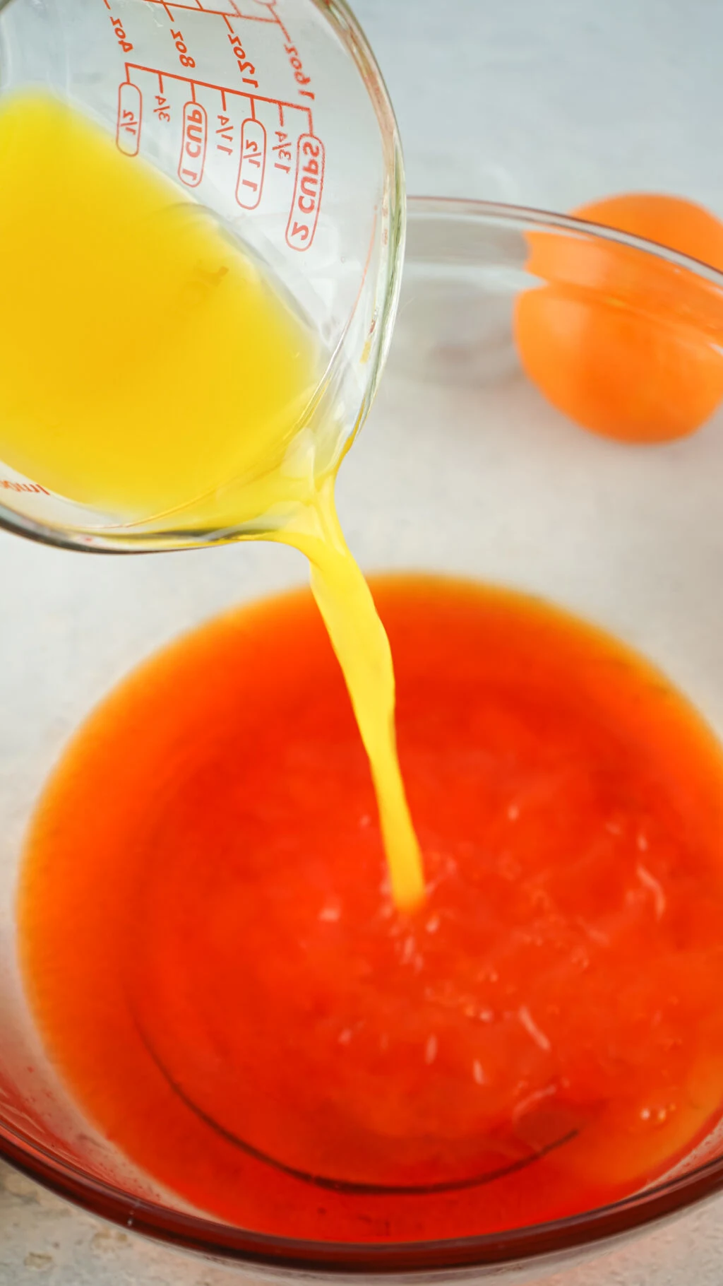 orange juice being poured into orange jello shot mixture in a clear glass bowl