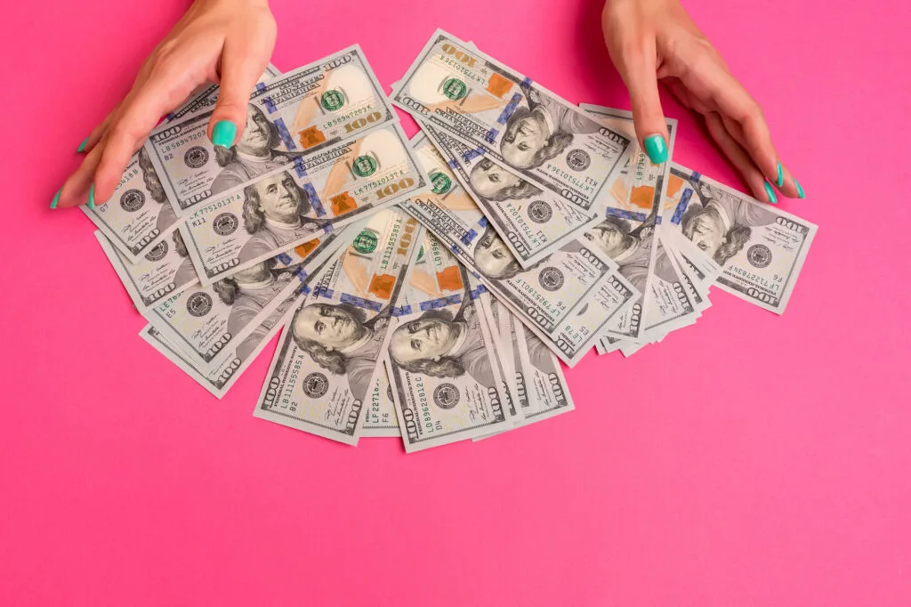 woman's hands with mint nails holding pile of money on hot pink table